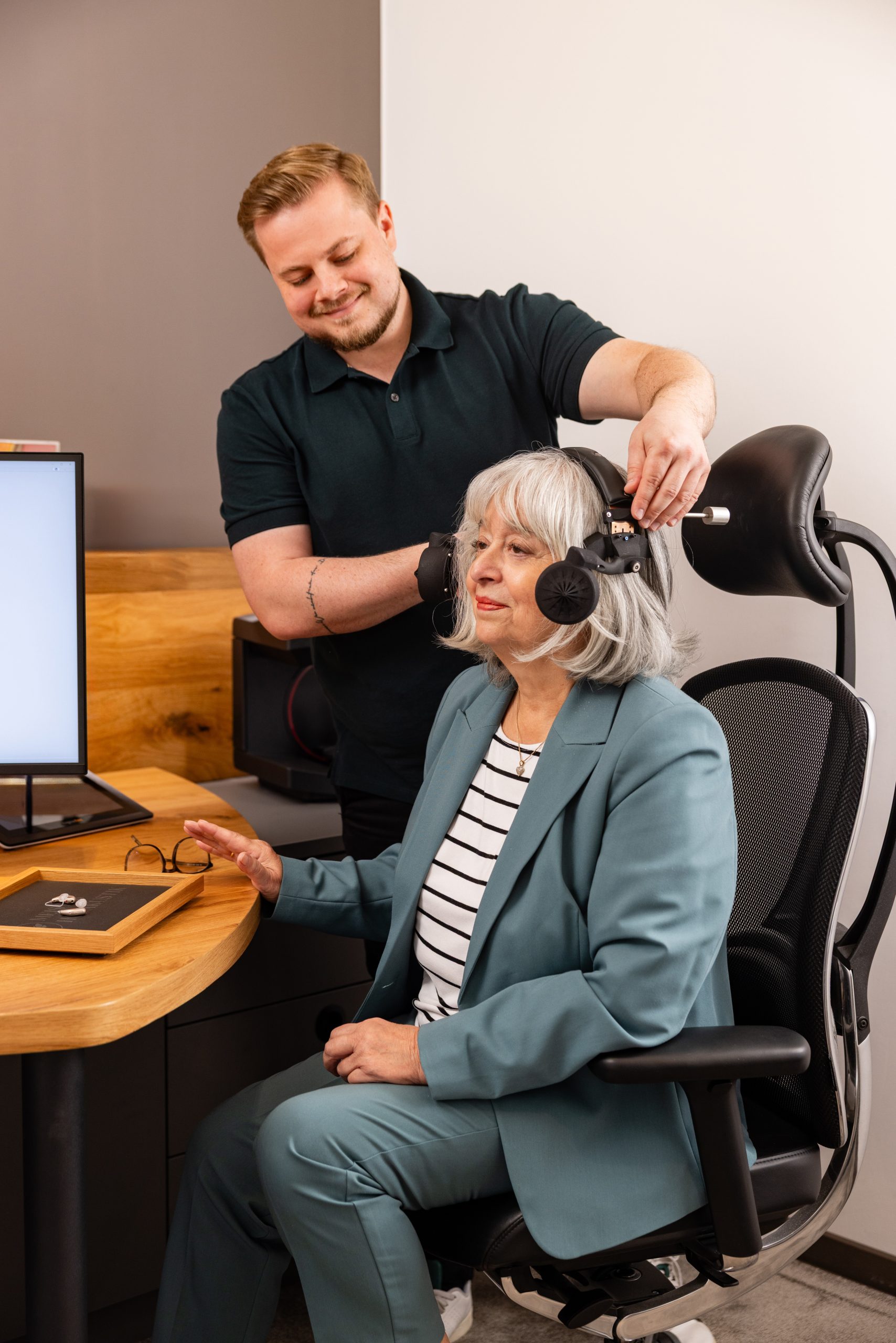 Hörakustikmeister besucht eine Kundin zuhause und setzt ihr einen Audiometriekopfhörer auf.